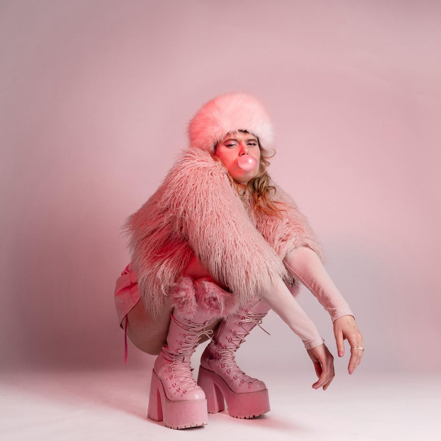 Een foto van een vrouw in roze kleding die gehurkt zit. Ze kijkt naar de camera en blaast een bel met kauwgom. Ze heeft ook een roze muts op. Ze draagt hoge plateauschoenen.